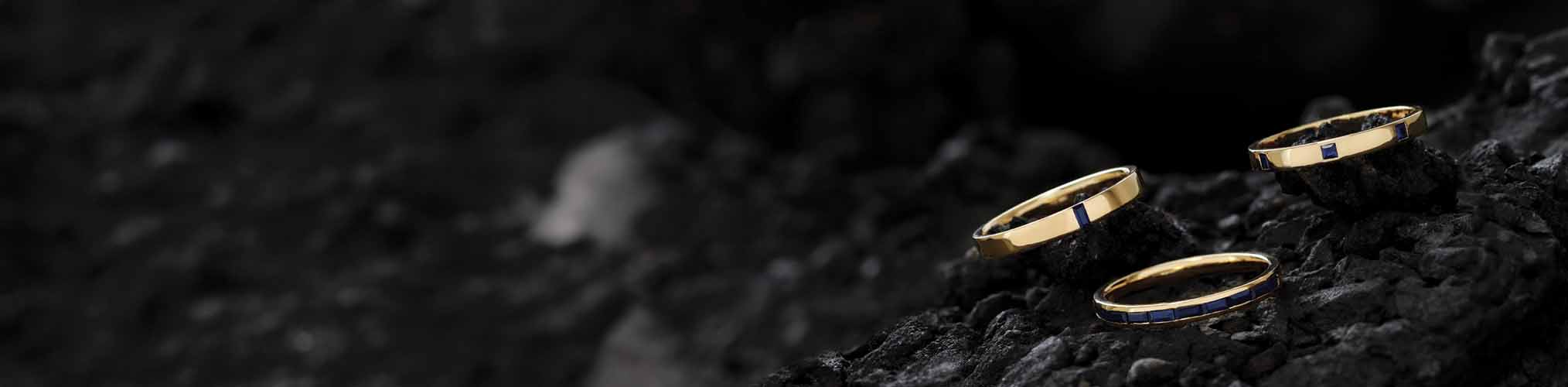 Three yellow gold men's wedding rings with blue sapphire inlays on textured black volcanic stone, modern jewellery