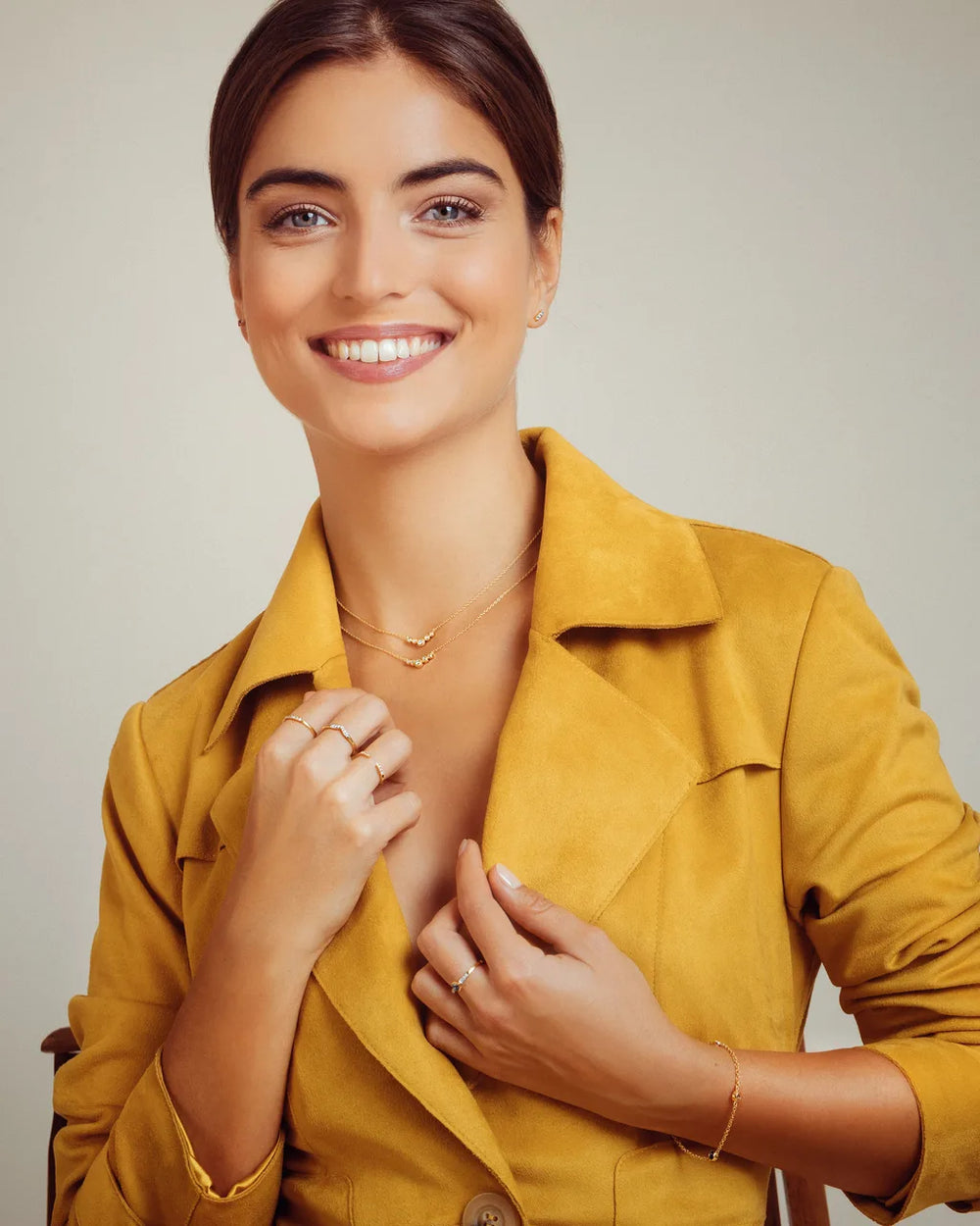 Yellow gold rings and bracelet worn by a woman in an ochre jacket on a beige background, Mayuri