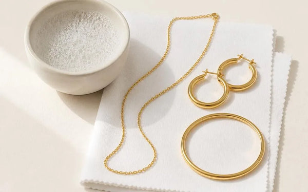 Gold-plated jewellery arranged on a microfibre cloth beside a bowl of soapy water, ready for gentle cleaning